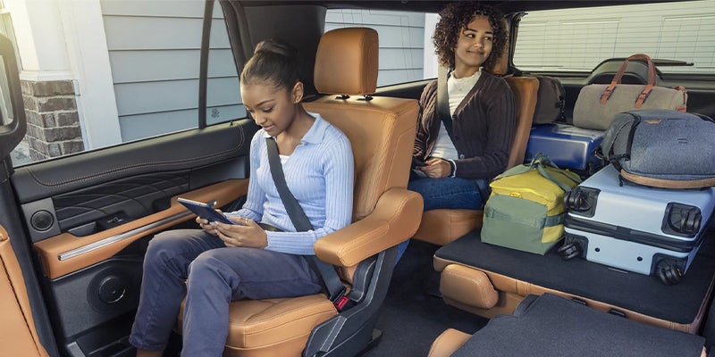 Two women seated in comfy tan leather seats inside a vehicle with plenty of luggage in the spacious back.