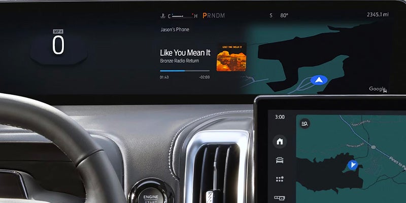 A car's digital dashboard displaying music, navigation, 0 MPH, and a smaller touchscreen with a map.