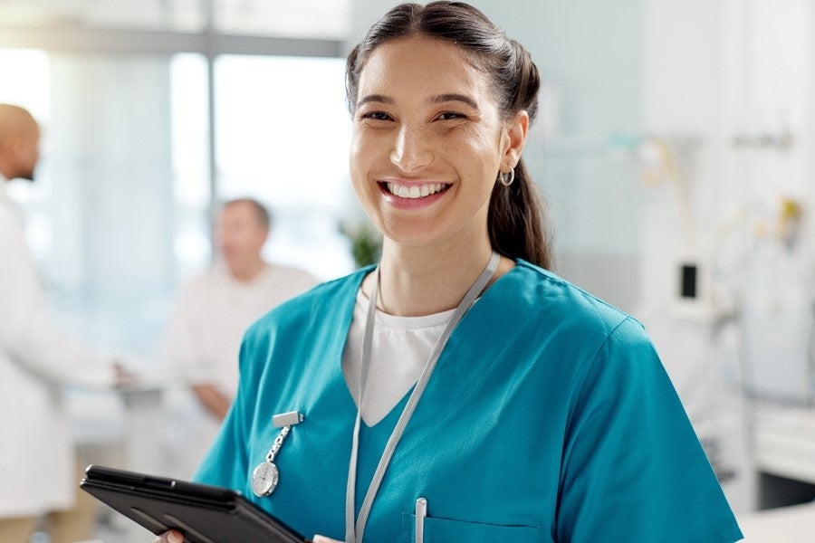 A smiling female healthcare professional in blue scrubs holds a tablet