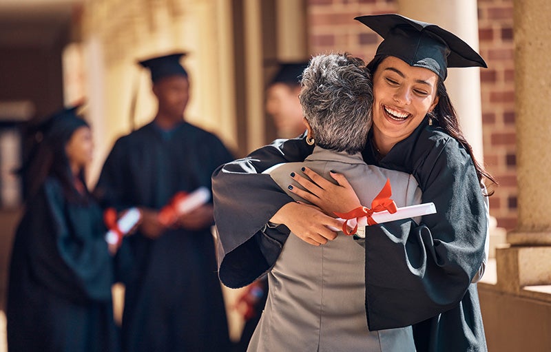 Smiling graduate in cap and gown hugging a woman while holding diploma